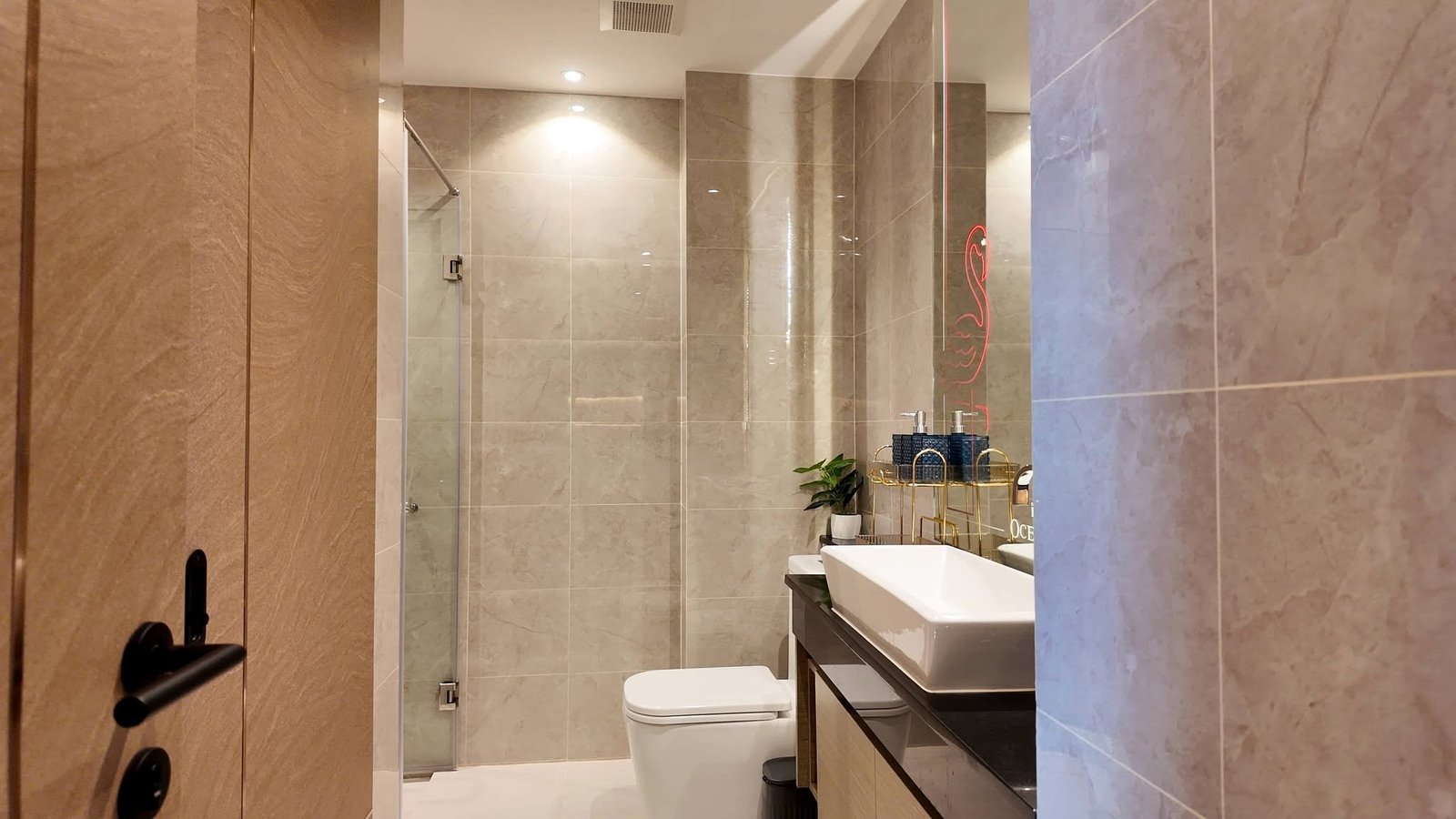 The_Riviera_Ocean_Drive_Pattaya_Dlc601_6 Modern bathroom with beige marble tiles, glass shower, white basin, and a sleek dark vanity; a potted plant sits by the sink.