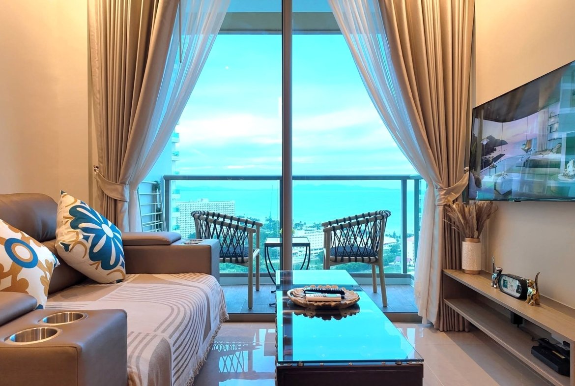 Living room with beige sofa and colorful floral cushions, glass coffee table, and balcony through sliding doors with sheer curtains opening to a blue sky and ocean view.