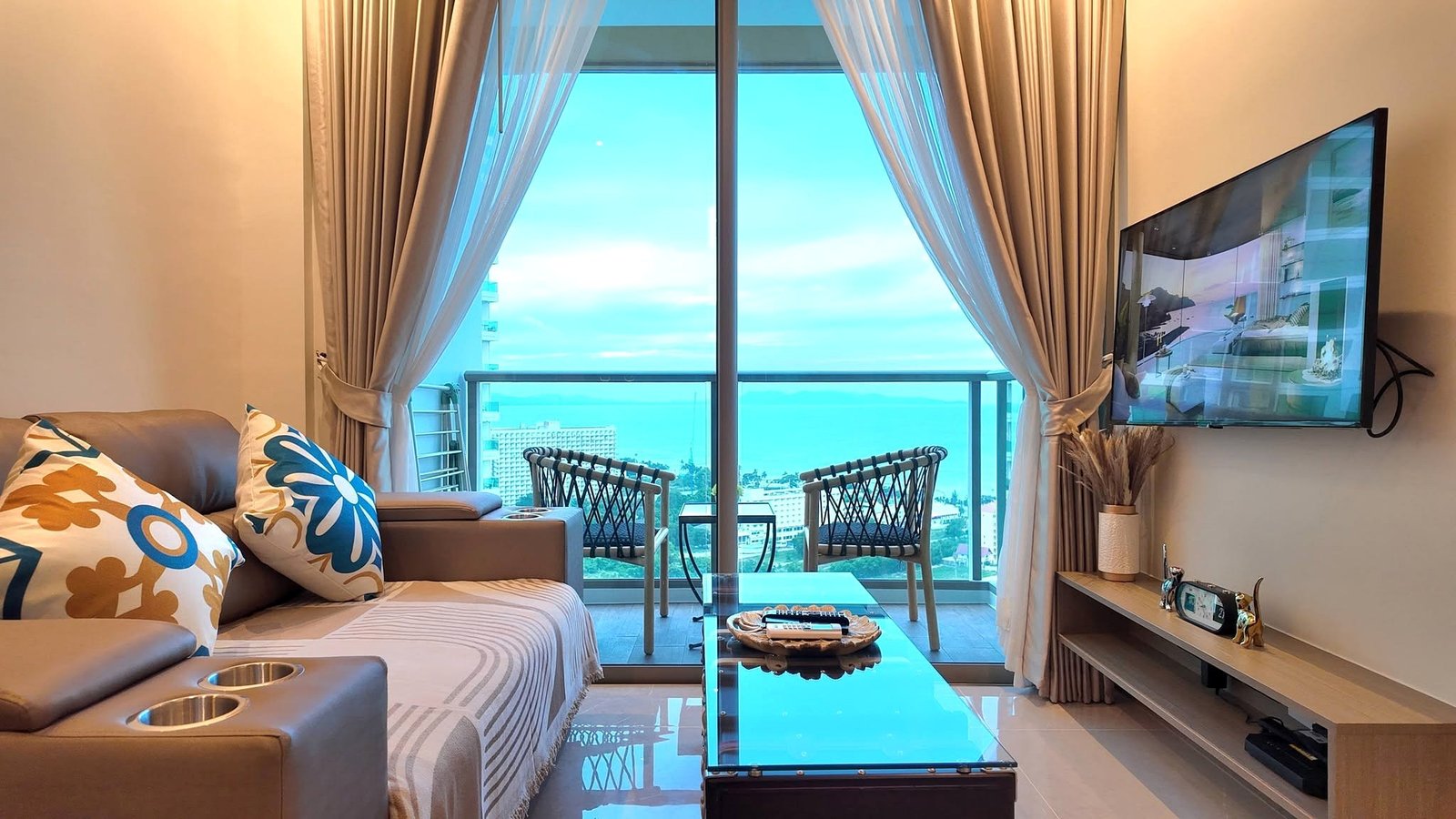 The_Riviera_Ocean_Drive_Pattaya_Dlc601_9 Living room with beige sofa and colorful floral cushions, glass coffee table, and balcony through sliding doors with sheer curtains opening to a blue sky and ocean view.