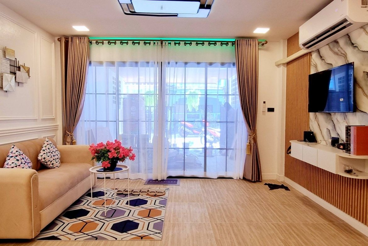 Living room with beige sectional sofa, geometric rug, and pink flowers on a round glass table by large glass doors with sheer curtains.