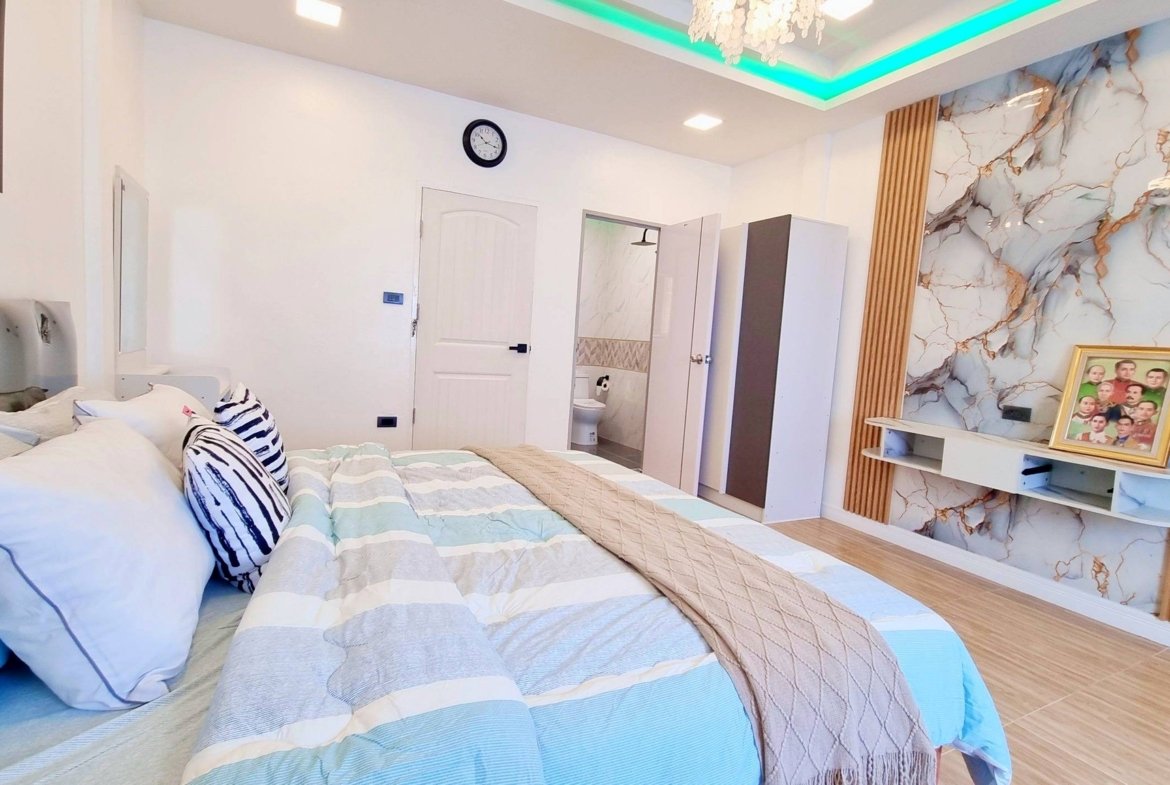 Modern bedroom with a large bed and blue-gray striped bedding, puffy pillows, and a marble accent wall with a framed family photo on a shelf nearby.