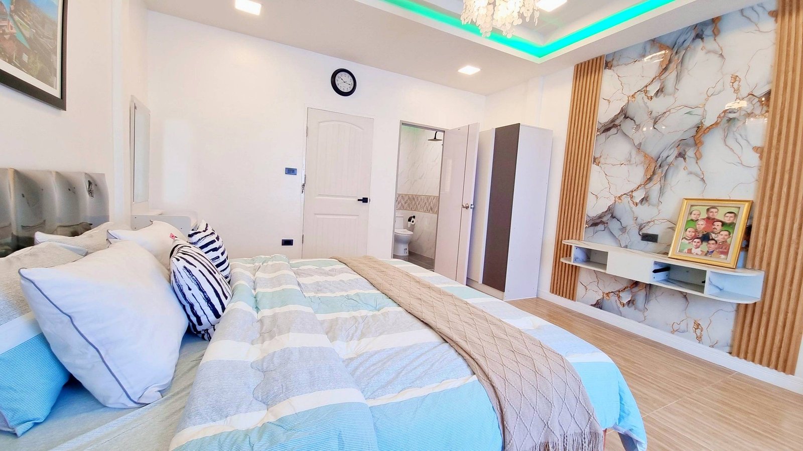 Town_home_Pattaya_Dlh551_15 Modern bedroom with a large bed and blue-gray striped bedding, puffy pillows, and a marble accent wall with a framed family photo on a shelf nearby.