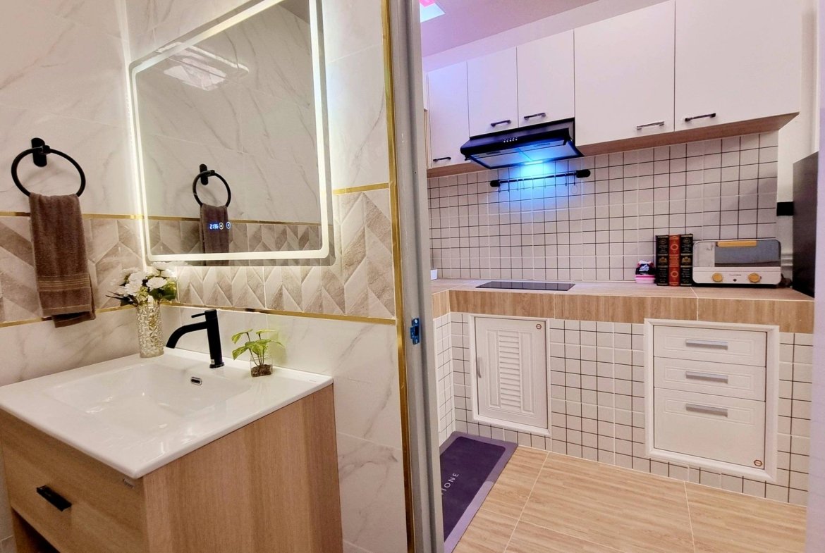 Modern bathroom vanity with white sink, black faucet, wood cabinet, and circular towel rings; glass partition leads to a tiled kitchen area.
