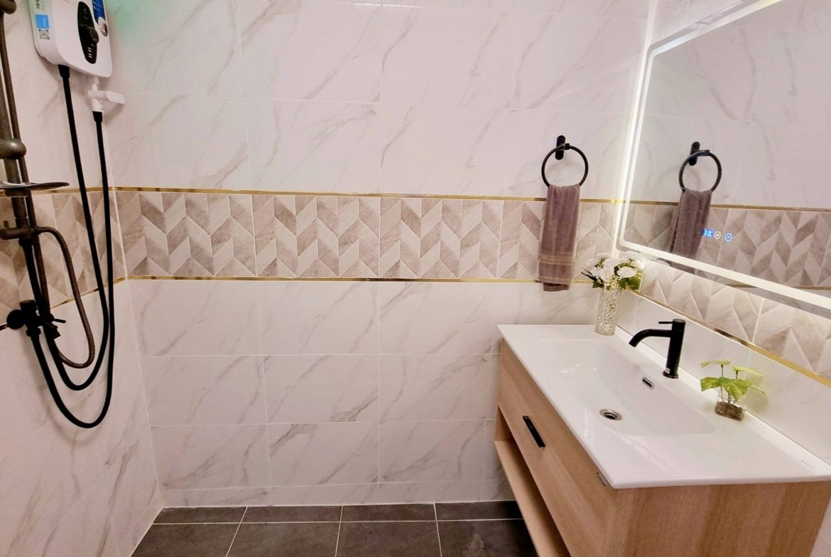 Modern bathroom showing a white tile wall with geometric accents, a wooden vanity with a white sink, and a large backlit mirror; two circular towel rings with gray towels on the wall; small vase with flowers on the counter; shower area with a handheld shower attachment on the left.