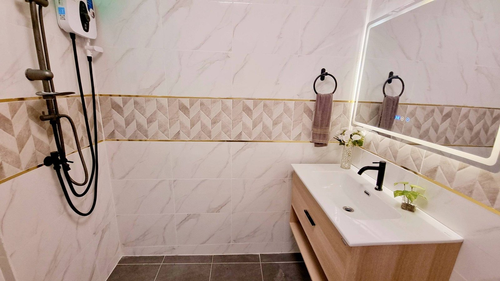 Town_home_Pattaya_Dlh551_17 Modern bathroom showing a white tile wall with geometric accents, a wooden vanity with a white sink, and a large backlit mirror; two circular towel rings with gray towels on the wall; small vase with flowers on the counter; shower area with a handheld shower attachment on the left.
