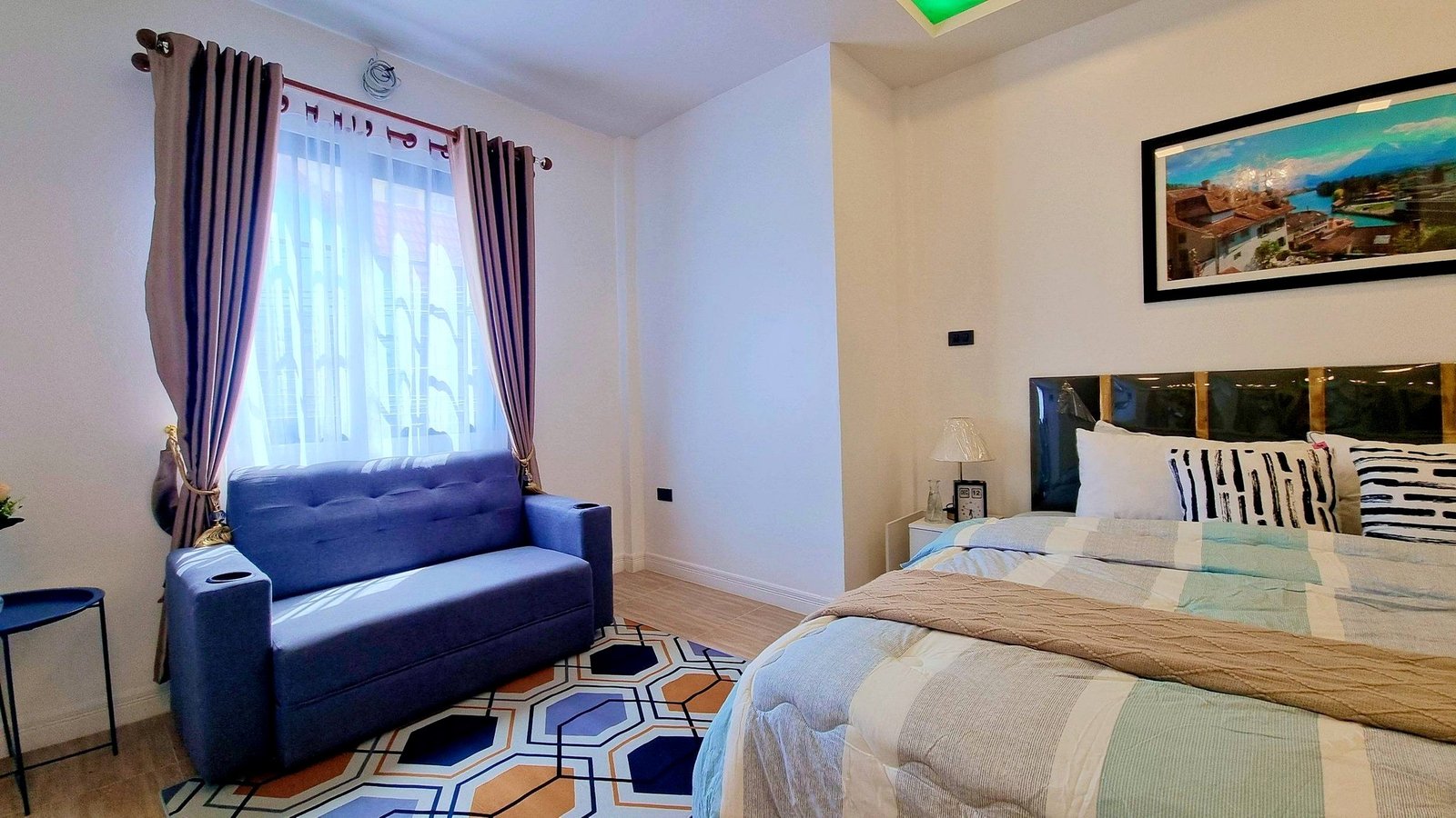 Town_home_Pattaya_Dlh551_6 Twin beds in a bright hotel room with a blue sofa by the window and sheer curtains.
