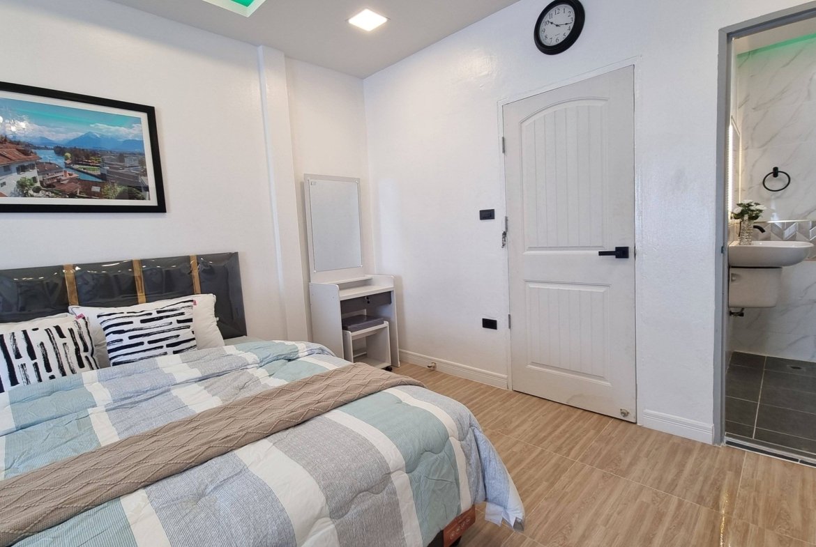 Bedroom with a striped quilt and decorative pillows, a framed landscape photo on the wall, and a door leading to a bathroom area on the right.