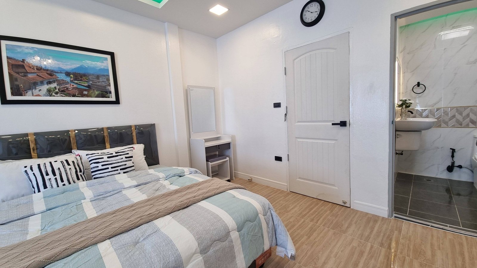 Town_home_Pattaya_Dlh551_7 Bedroom with a striped quilt and decorative pillows, a framed landscape photo on the wall, and a door leading to a bathroom area on the right.
