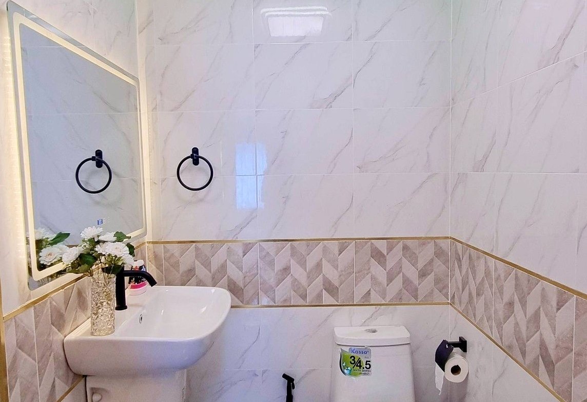 Small modern bathroom with white sink, wall mirror, black towel rings, and a white toilet under a green LED strip along marble-tiled walls.