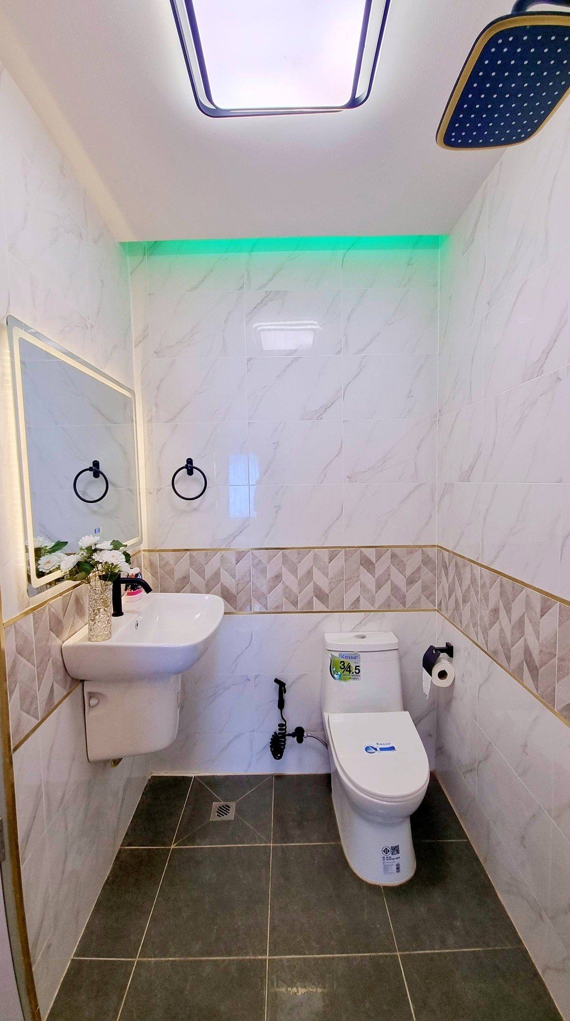 Town_home_Pattaya_Dlh551_8 Small modern bathroom with white sink, wall mirror, black towel rings, and a white toilet under a green LED strip along marble-tiled walls.
