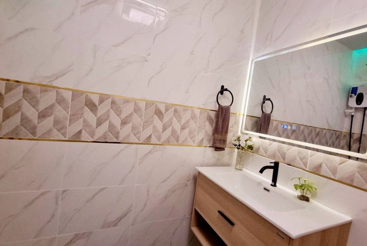 Modern bathroom vanity with rectangular backlit mirror, white sink, black faucet, wooden cabinet, and brown towel rings on marble-tiled walls.