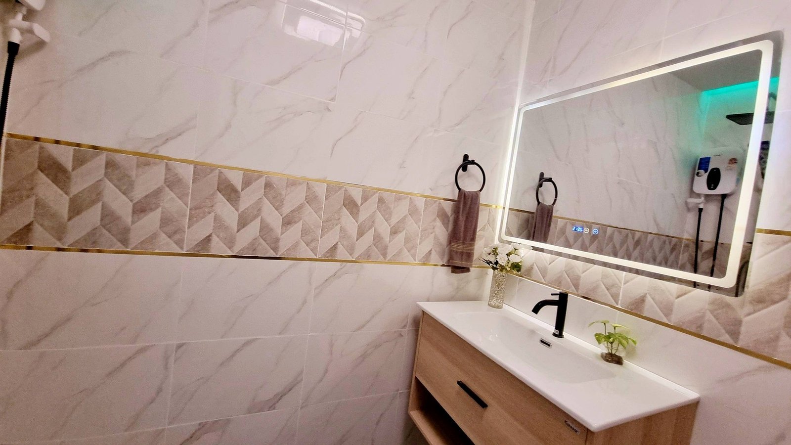 Town_home_Pattaya_Dlh551_9 Modern bathroom vanity with rectangular backlit mirror, white sink, black faucet, wooden cabinet, and brown towel rings on marble-tiled walls.