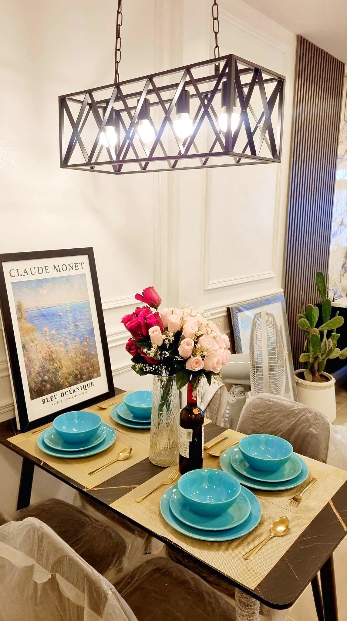 Townhome_South_Pattaya_Dlh581_10 Dining table set with turquoise bowls, gold utensils, and a pink bouquet centerpiece under a geometric chandelier; Monet poster leaning on the wall in a stylish dining room.