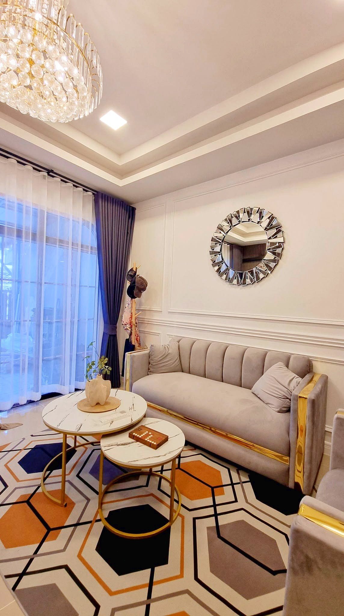 Townhome_South_Pattaya_Dlh581_13 Elegant living room with a crystal chandelier, light beige sofa, and marble-top nesting tables on a geometric orange, gray, and black rug.