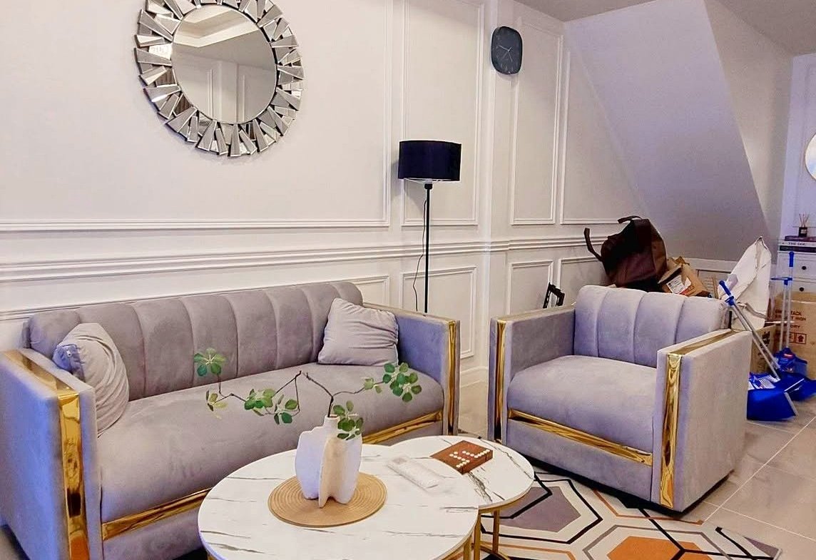 Modern living room with gray velvet sofas and gold frames, white paneled walls, and a geometric orange-gray rug under round marble-topped coffee tables.