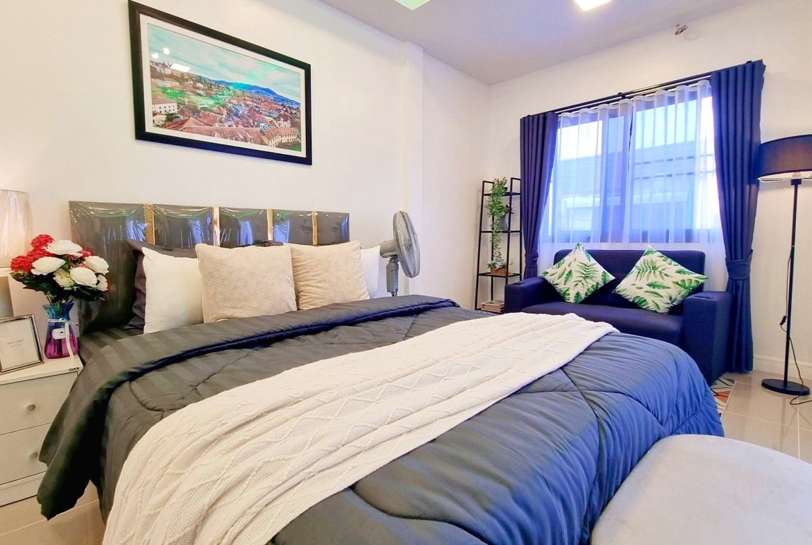 Bedroom with blue bedding, multiple pillows, and a white nightstand holding a vase of red and white flowers next to a framed photo.