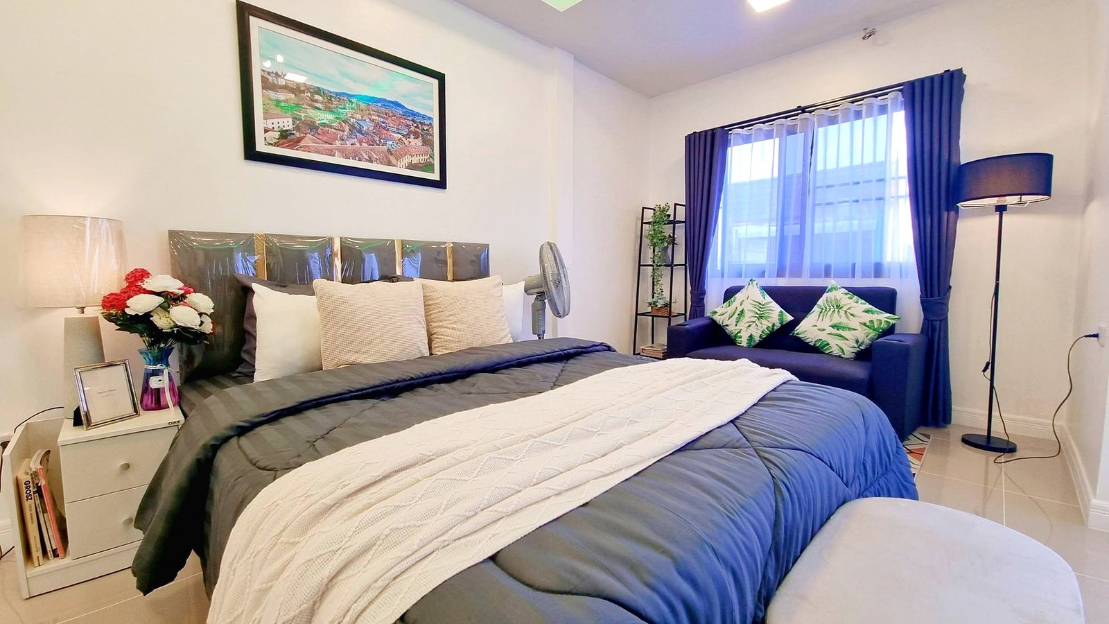 Townhome_South_Pattaya_Dlh581_16 Bedroom with blue bedding, multiple pillows, and a white nightstand holding a vase of red and white flowers next to a framed photo.