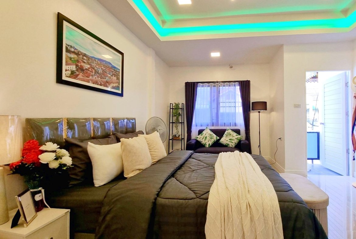 Cozy living area with a large dark gray bed/sofa, cream pillows, and a white side table bearing flowers, plus a framed cityscape on the wall and a window with dark curtains.