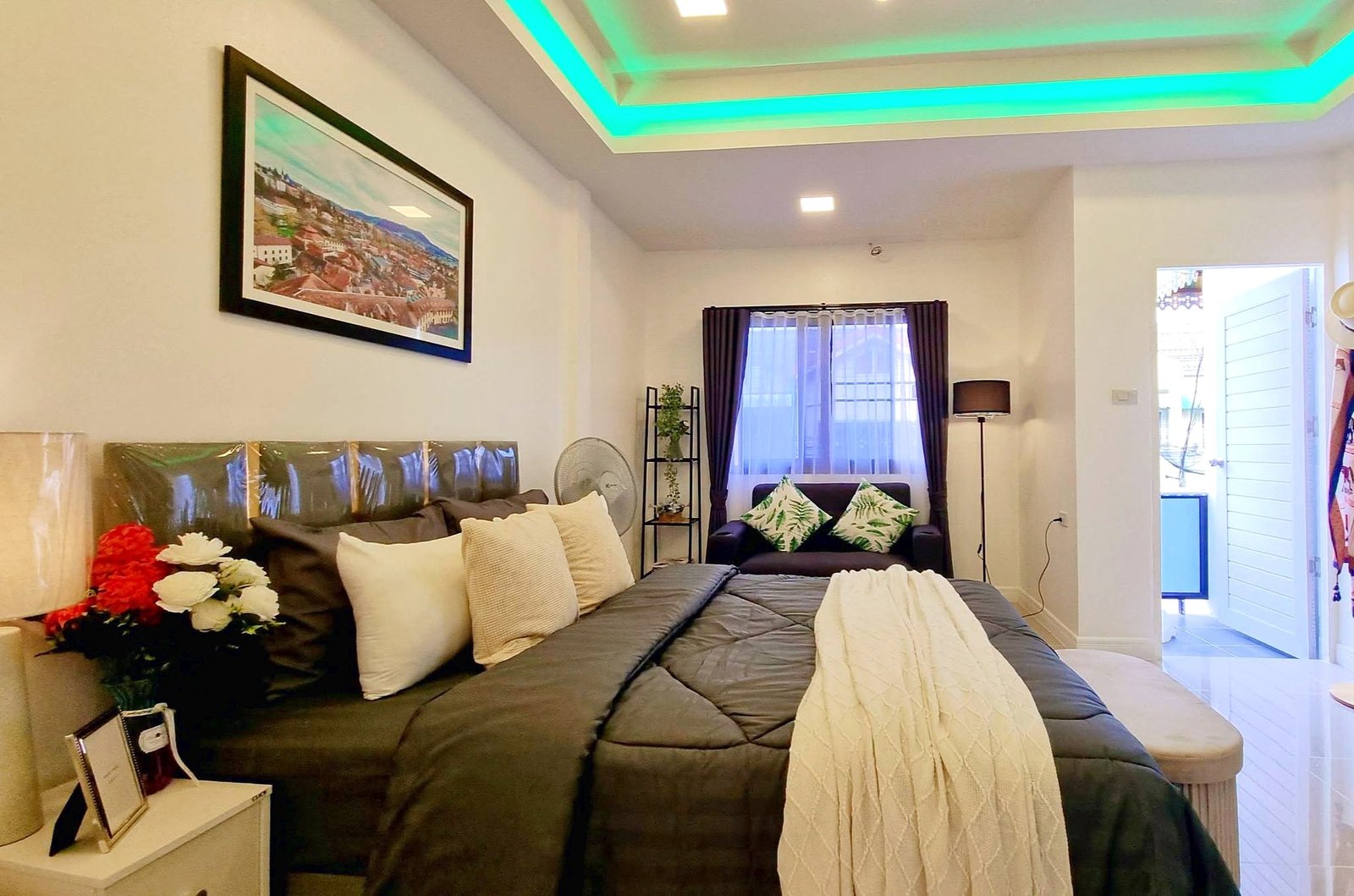 Townhome_South_Pattaya_Dlh581_17 Cozy living area with a large dark gray bed/sofa, cream pillows, and a white side table bearing flowers, plus a framed cityscape on the wall and a window with dark curtains.
