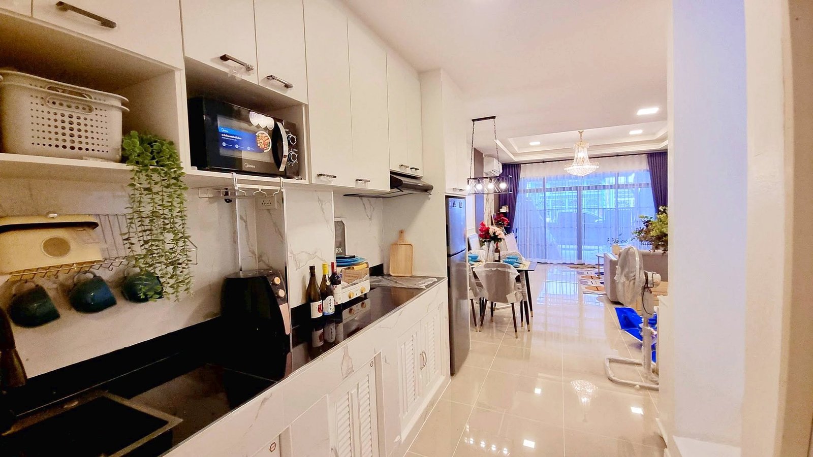 Townhome_South_Pattaya_Dlh581_20 Modern white kitchen with microwave above the counter, hanging plant, and dish rack, opening to the dining area with a chandelier.
