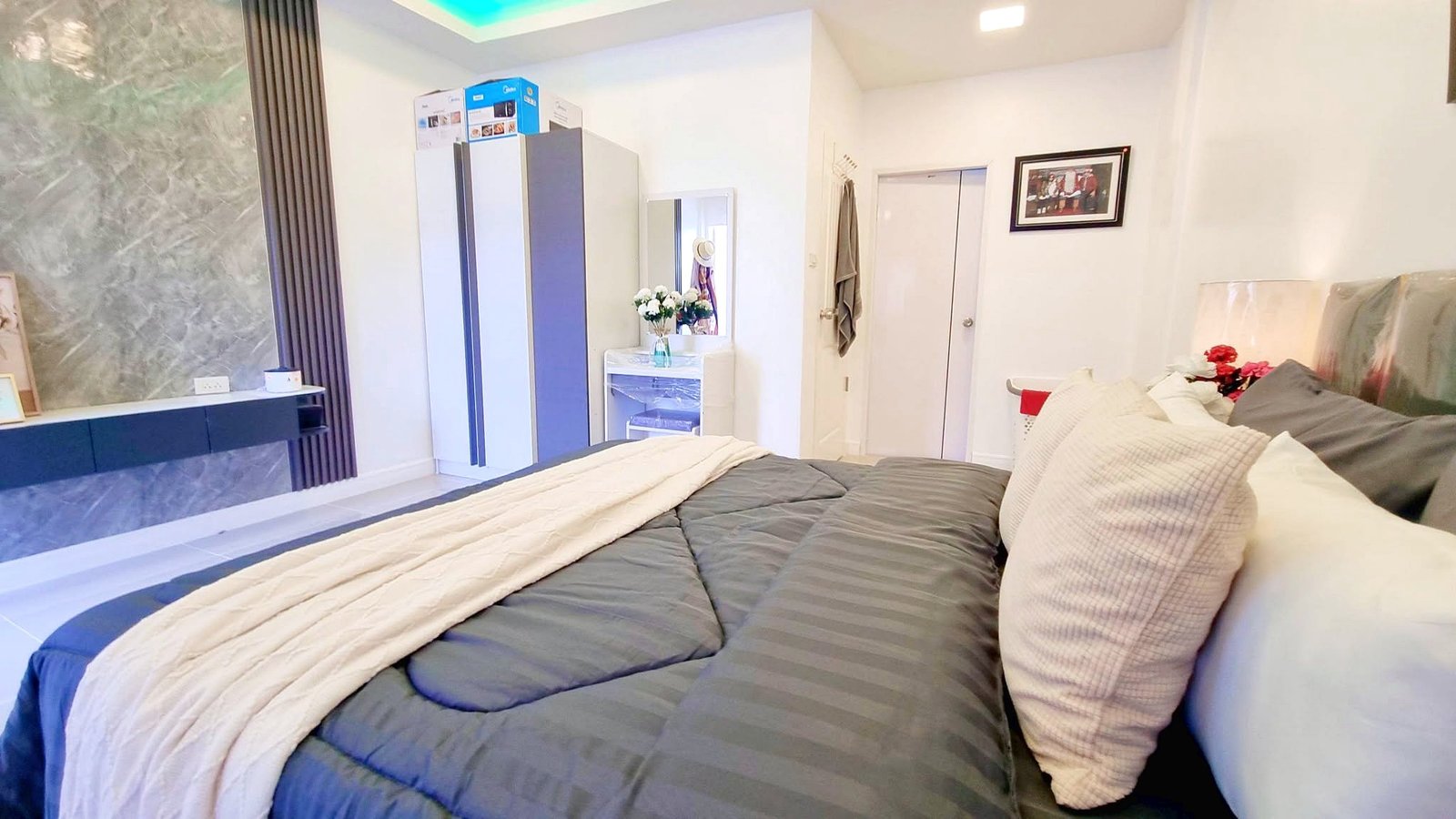 Townhome_South_Pattaya_Dlh581_4 Large bed in a modern bedroom with gray striped bedding, a beige throw, and white pillows; marble accent wall and dresser with a mirror in the background.