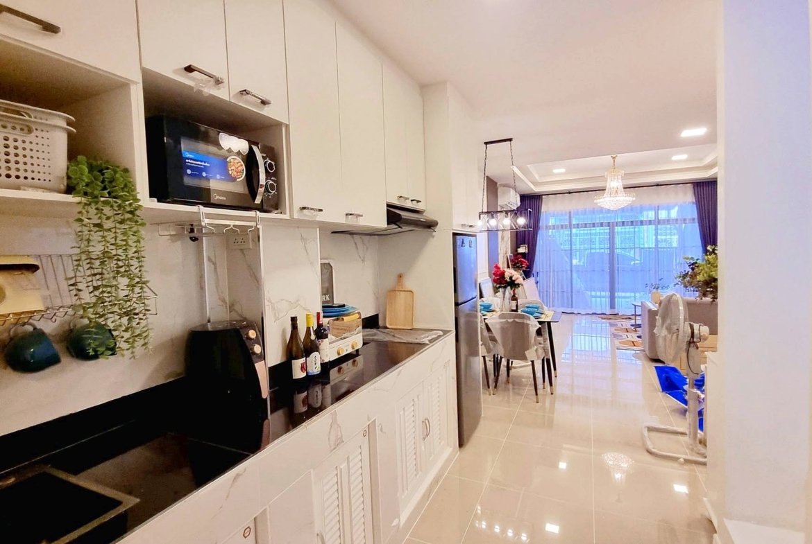Bright modern kitchen with white cabinets, a microwave built in above the counter, and hanging greenery on an open shelf leading to a dining area with a chandelier and large windows.