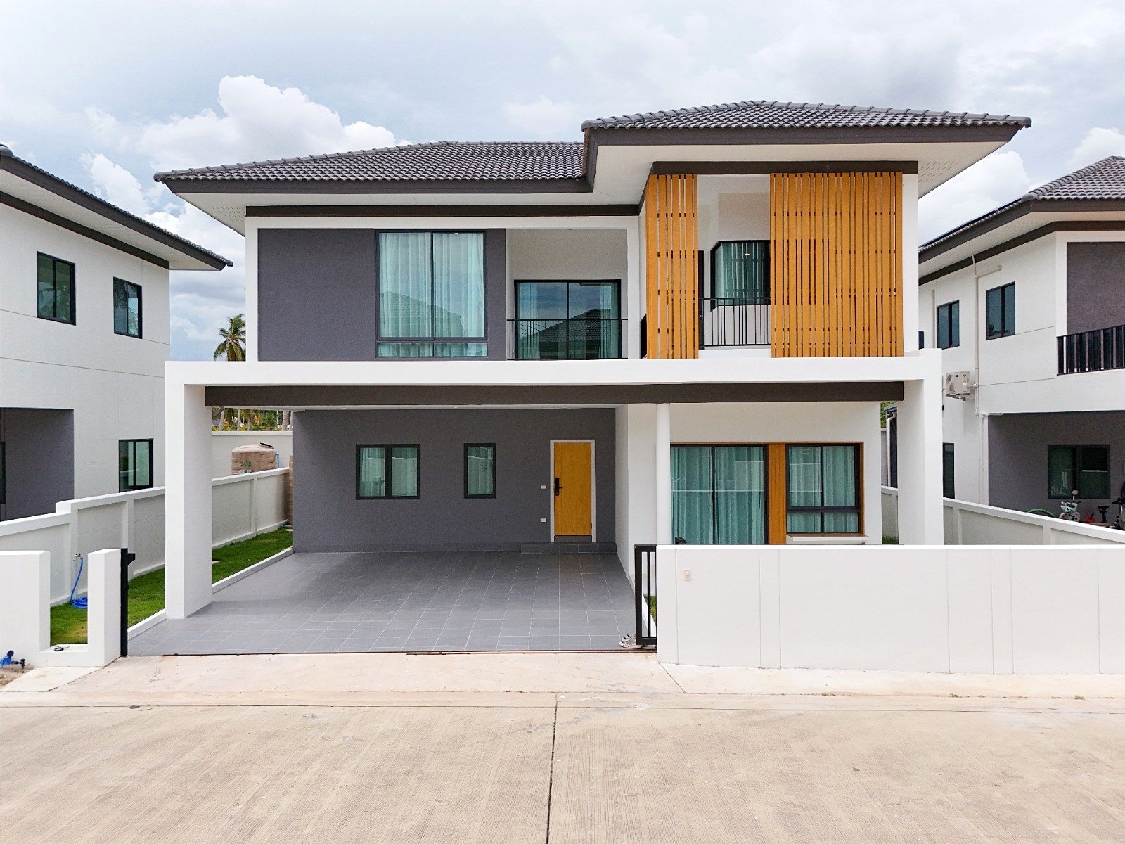 Tropical_Village_3_Pattaya_Dlh453 Modern two-story house with grey and white facade, wooden vertical slats, and a covered carport; bright yellow front door visible.