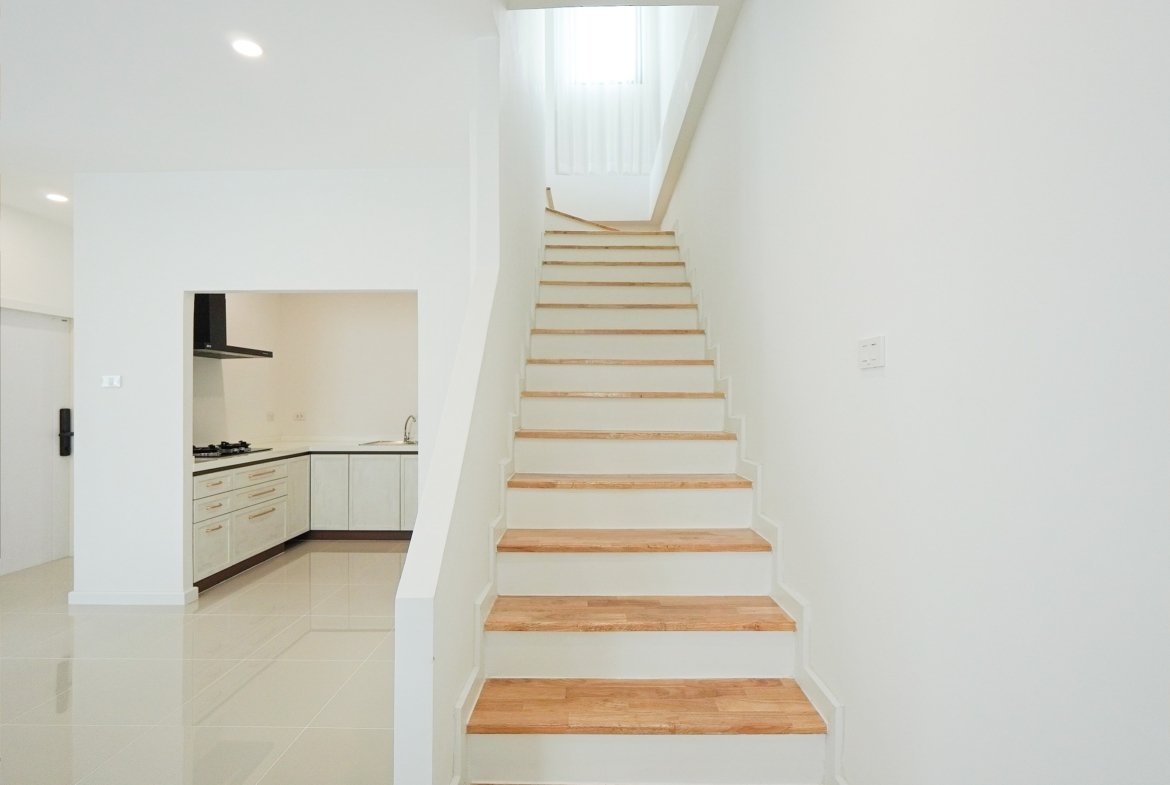 White staircase with wooden treads leading up to a bright landing, kitchen opening on the left in a minimalist white interior.