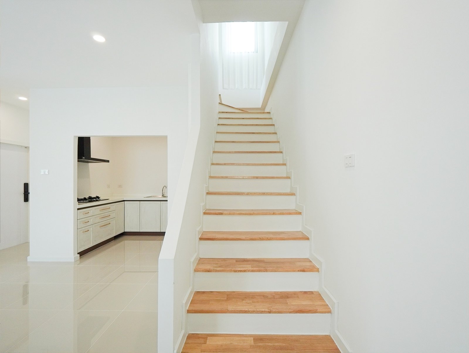 Tropical_Village_3_Pattaya_Dlh453_10 White staircase with wooden treads leading up to a bright landing, kitchen opening on the left in a minimalist white interior.
