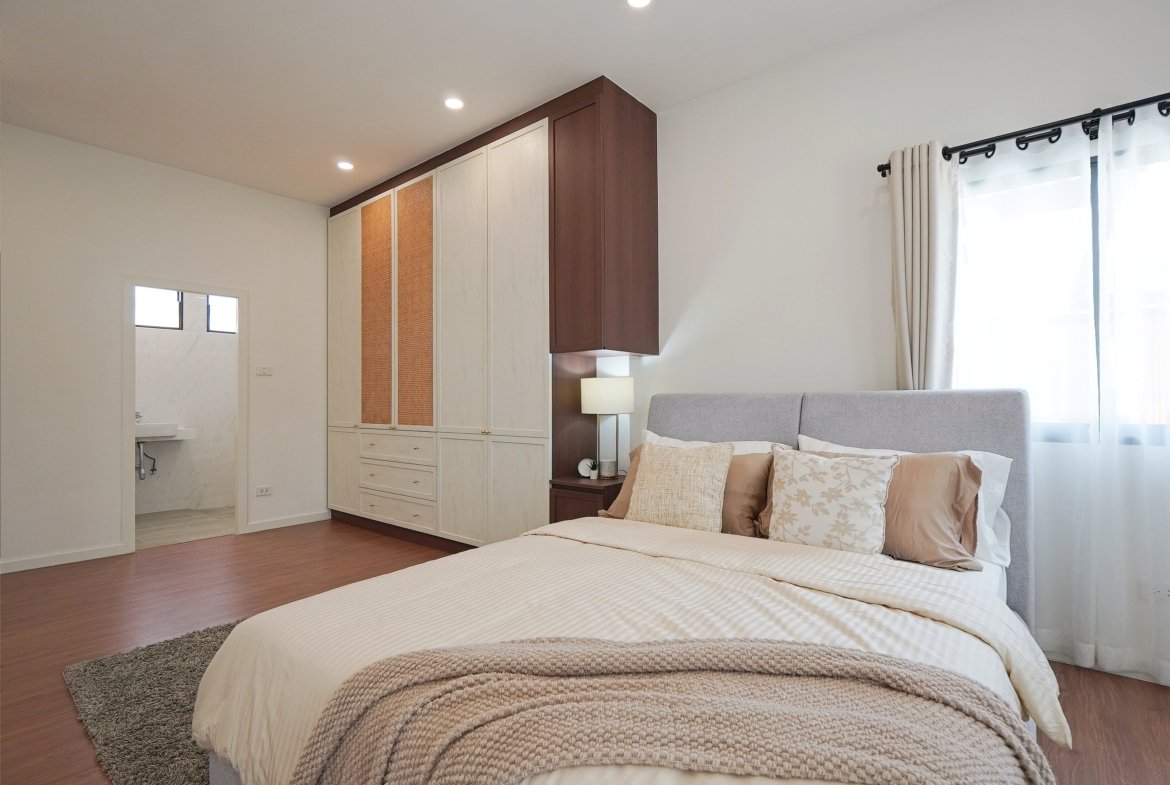 Modern bedroom with a large beige wardrobe, brown accents, and a gray upholstered bed. A bedside lamp sits beside the bed, and a window with white curtains is on the right.