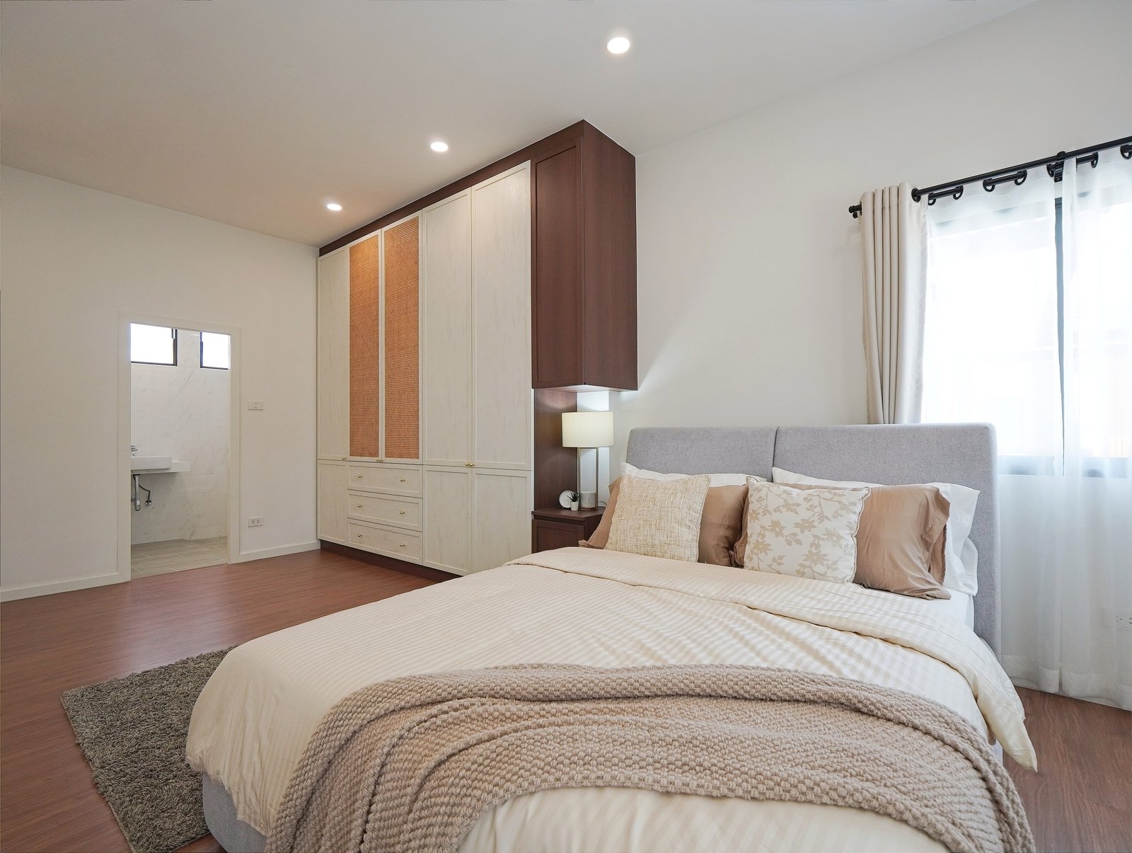 Tropical_Village_3_Pattaya_Dlh453_15 Modern bedroom with a large beige wardrobe, brown accents, and a gray upholstered bed. A bedside lamp sits beside the bed, and a window with white curtains is on the right.
