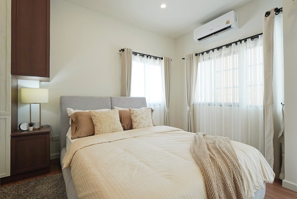 Bright hotel-style bedroom with a neatly made bed, beige bedding, and decorative throw pillows under an wall-mounted air conditioner. The room has sheer white curtains and a dark wood cabinet on the left.