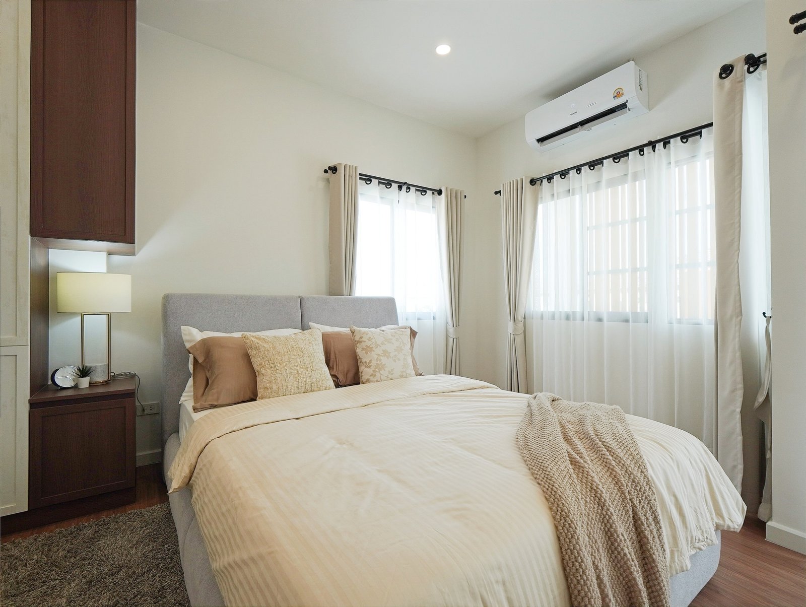 Tropical_Village_3_Pattaya_Dlh453_17 Bright hotel-style bedroom with a neatly made bed, beige bedding, and decorative throw pillows under an wall-mounted air conditioner. The room has sheer white curtains and a dark wood cabinet on the left.