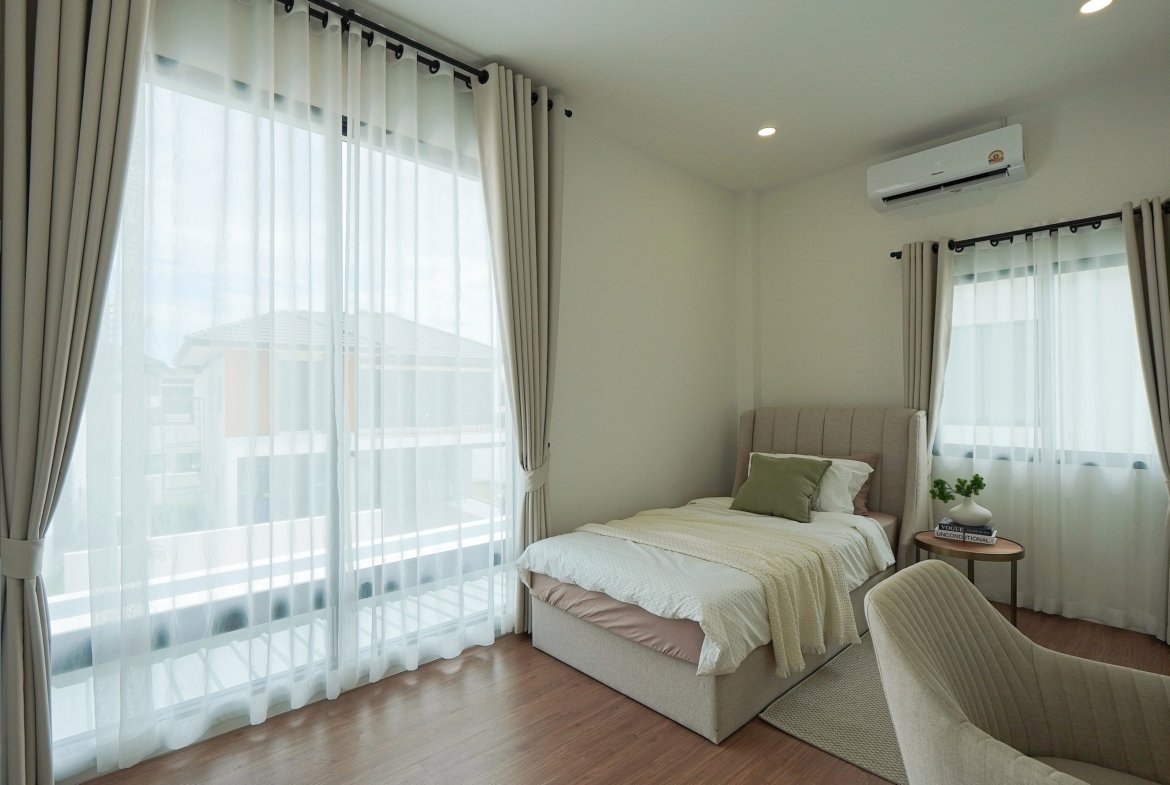 Bright bedroom with a gray upholstered bed, sheer white curtains, and a wall-mounted air conditioner letting in daylight from tall windows.