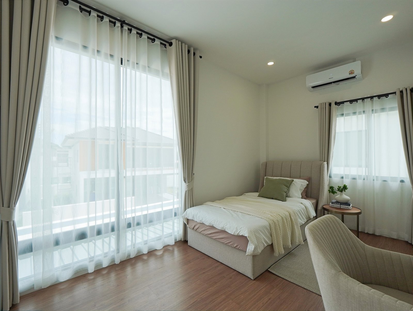 Tropical_Village_3_Pattaya_Dlh453_19 Bright bedroom with a gray upholstered bed, sheer white curtains, and a wall-mounted air conditioner letting in daylight from tall windows.