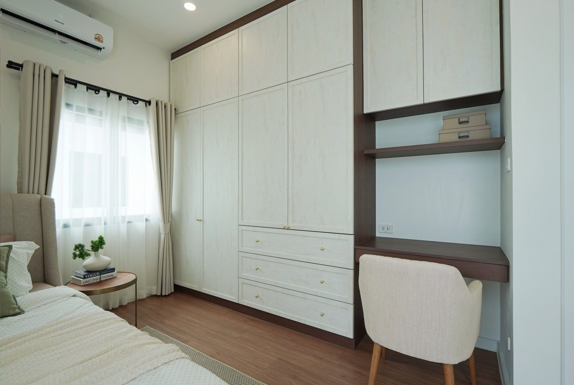 Bedroom with white built-in wardrobes and dark wood trim, a desk area with shelves and small boxes, a beige upholstered chair, and a bed with white bedding beside a window with sheer curtains.