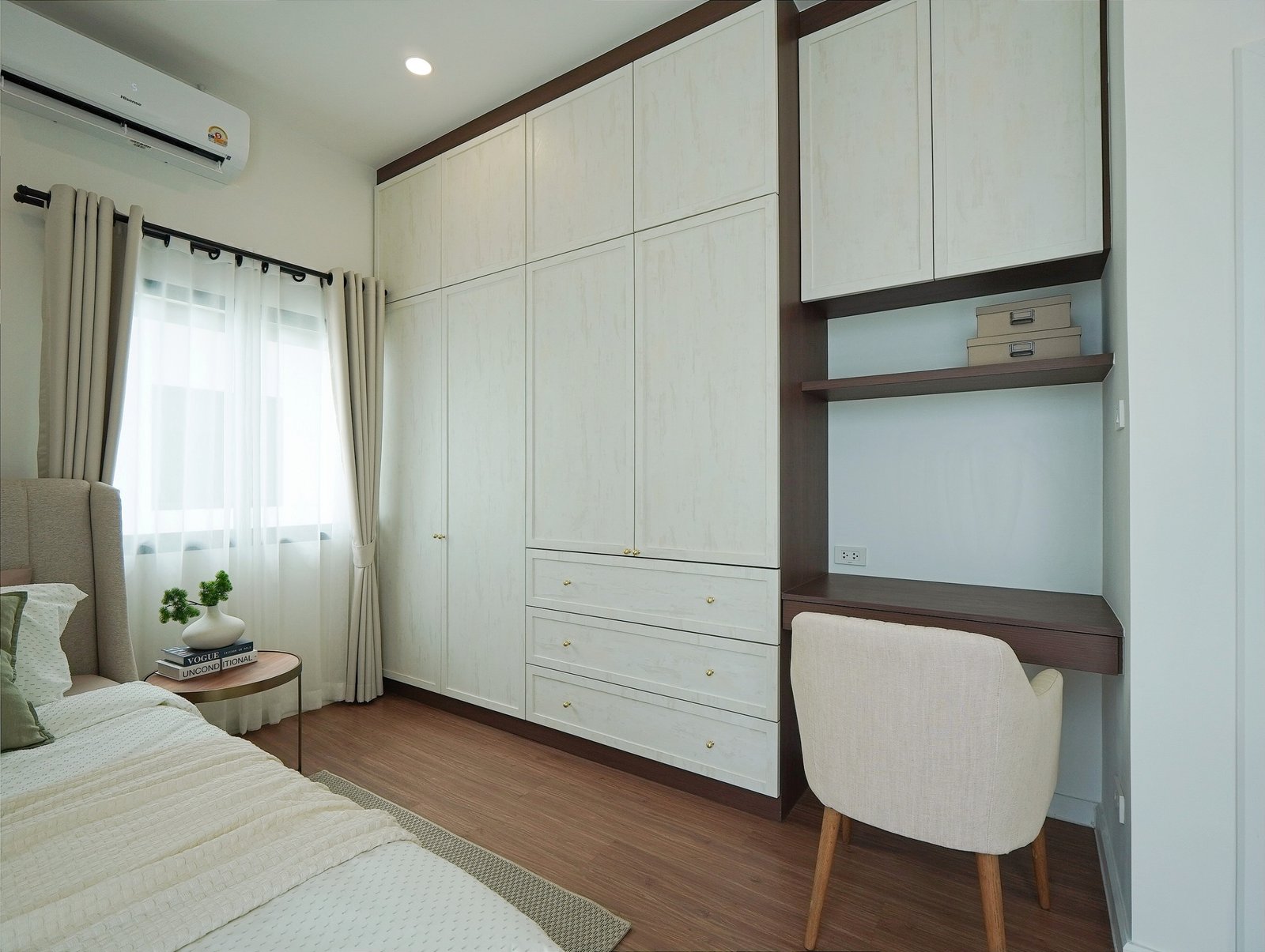 Tropical_Village_3_Pattaya_Dlh453_20 Bedroom with white built-in wardrobes and dark wood trim, a desk area with shelves and small boxes, a beige upholstered chair, and a bed with white bedding beside a window with sheer curtains.