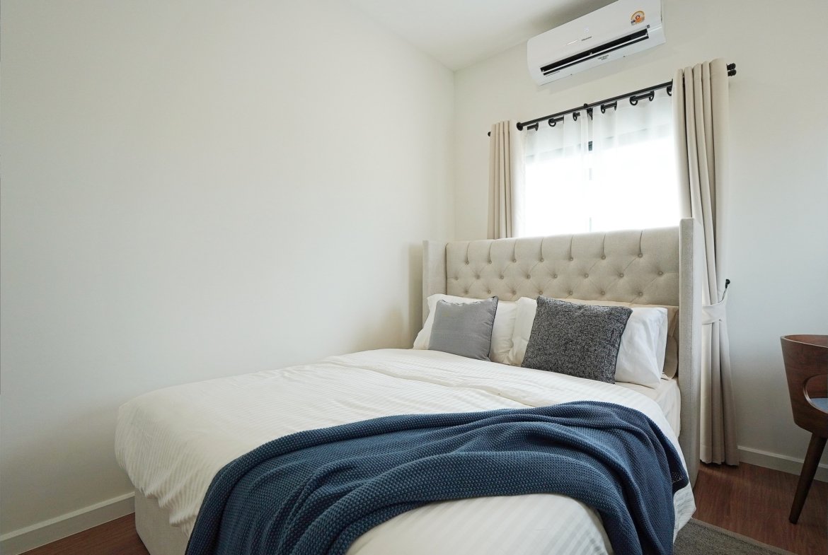Neatly made bedroom with a tufted headboard bed, white bedding, and a navy blue throw blanket near the foot of the bed, window with beige curtains.
