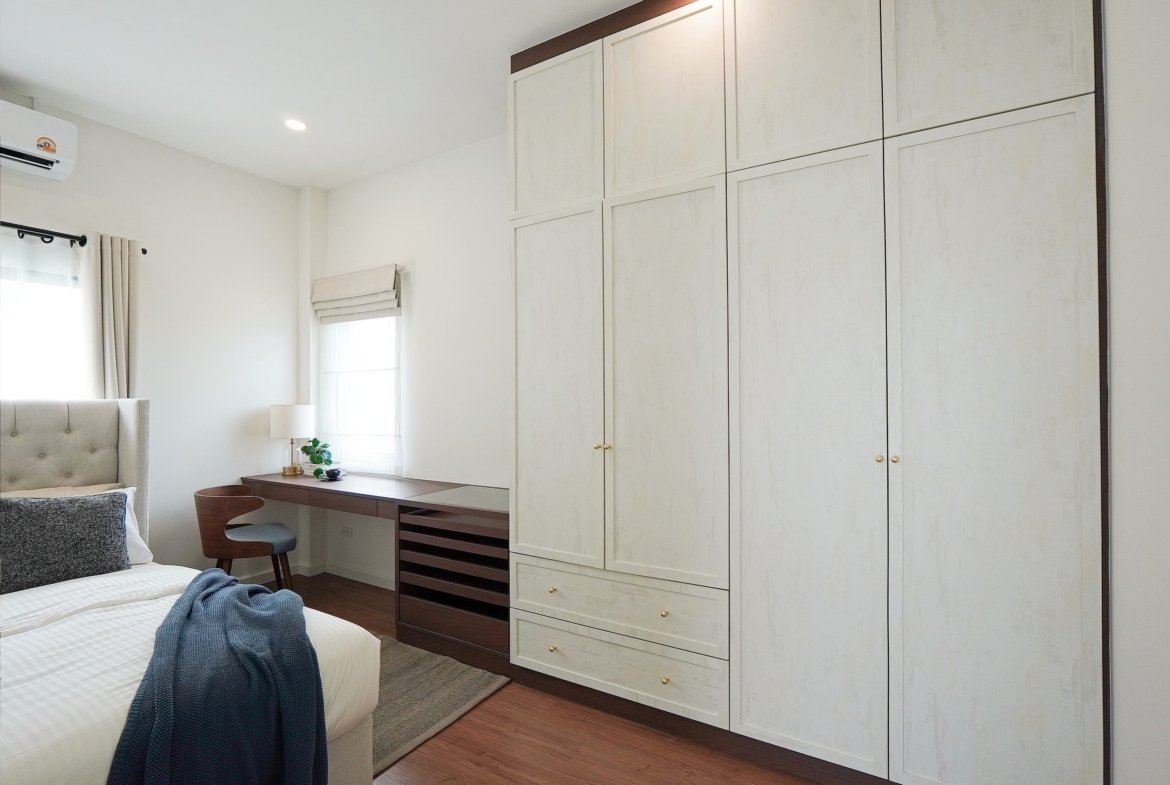 Bright bedroom with tall white paneled wardrobes along the right, a bed on the left, and a wooden desk by a window.