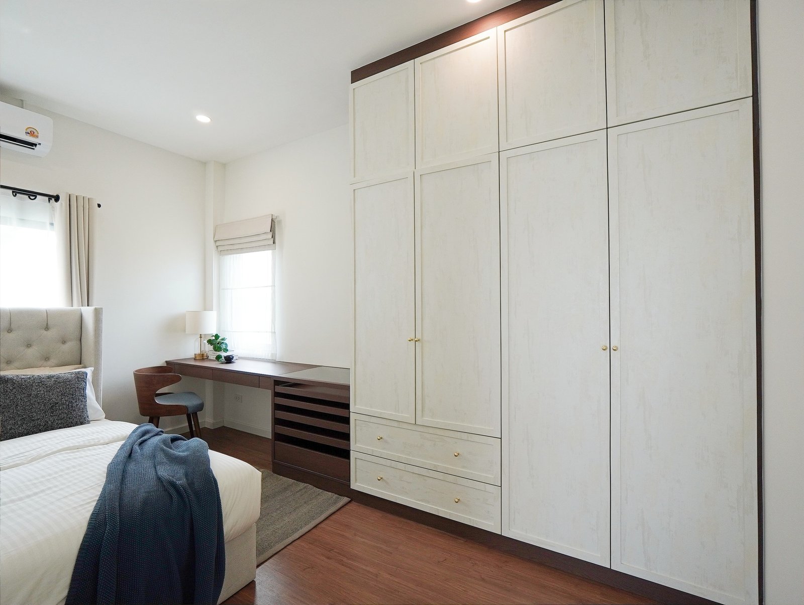 Tropical_Village_3_Pattaya_Dlh453_22 Bright bedroom with tall white paneled wardrobes along the right, a bed on the left, and a wooden desk by a window.