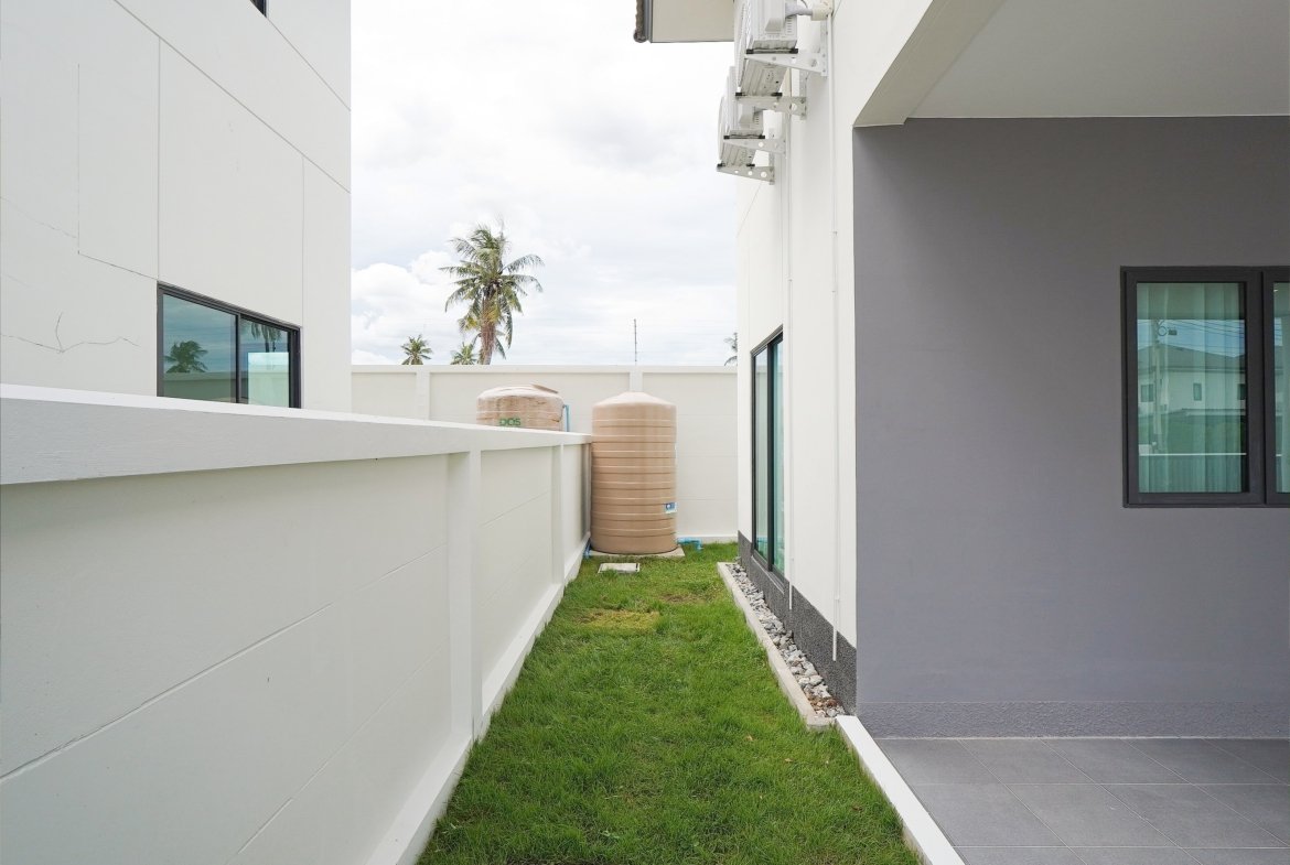 Narrow alley between white building walls with a grass strip, water tanks, and palm trees visible in the background.