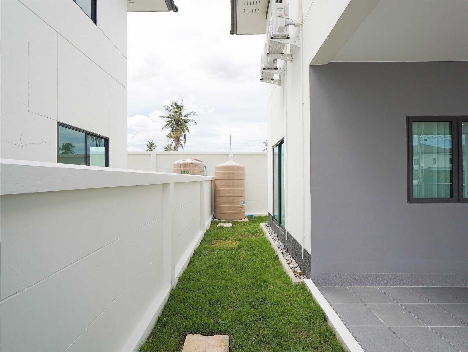 Tropical_Village_3_Pattaya_Dlh453_23 Narrow alley between white building walls with a grass strip, water tanks, and palm trees visible in the background.