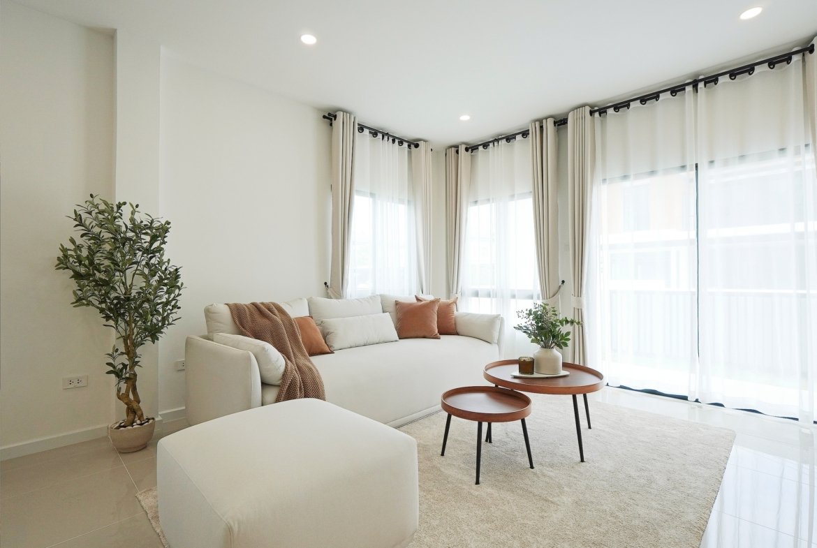 Bright living room with a white sofa, beige rug, and potted plant by large windows with sheer white curtains.
