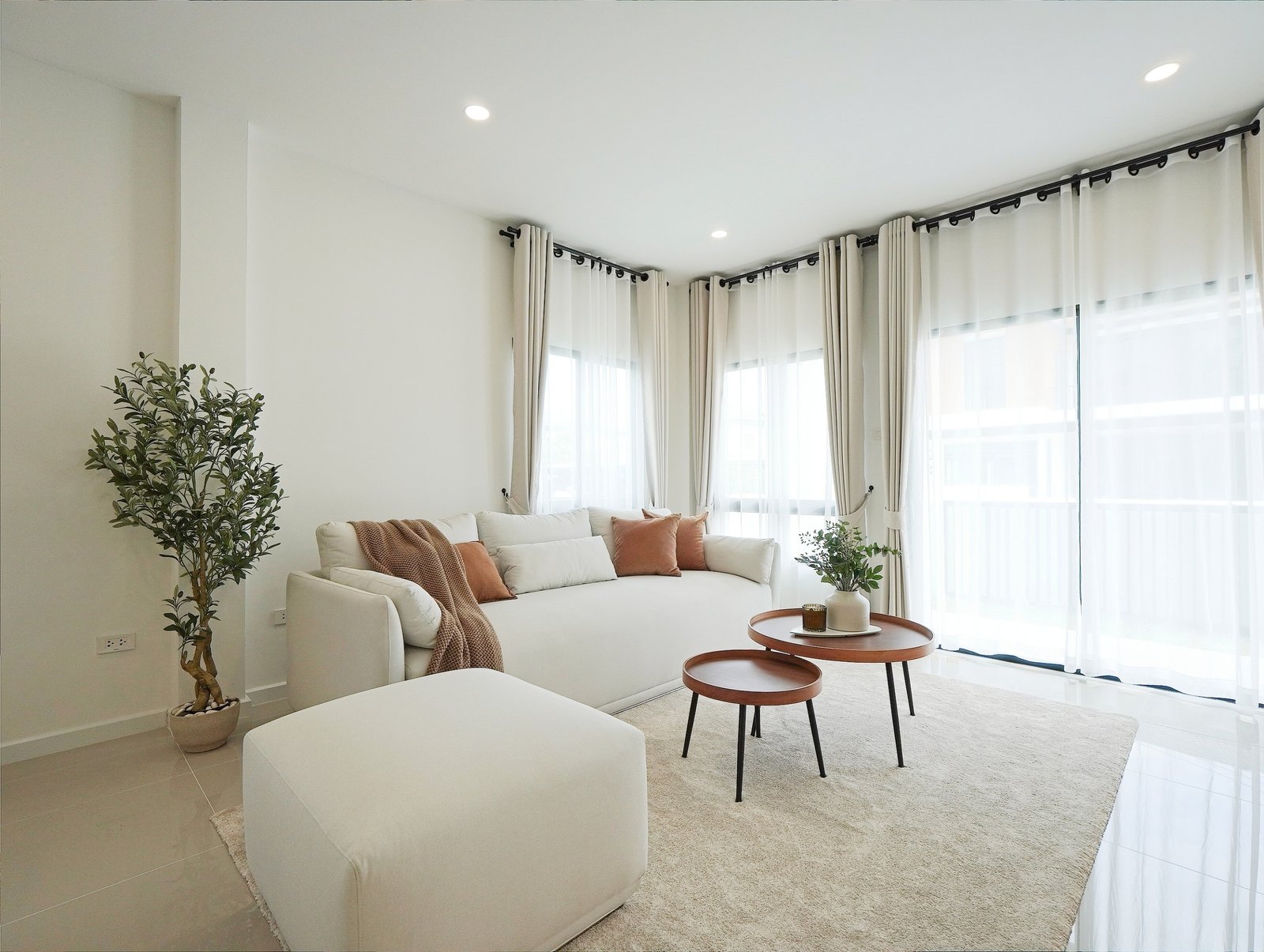 Tropical_Village_3_Pattaya_Dlh453_3 Bright living room with a white sofa, beige rug, and potted plant by large windows with sheer white curtains.