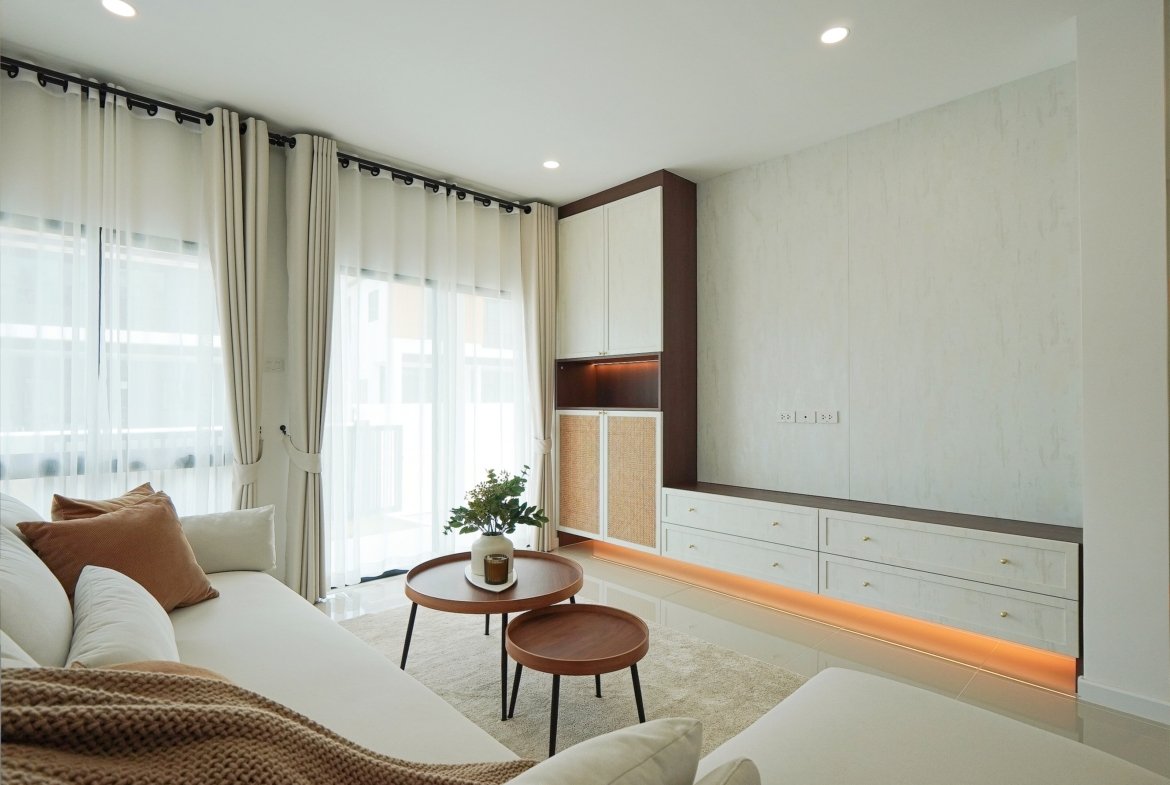 Bright living room with sheer curtains, white walls, and built-in cabinets; white sofa with brown throw and round wooden tables.