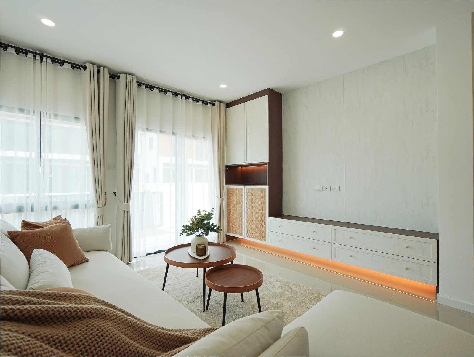 Tropical_Village_3_Pattaya_Dlh453_4 Bright living room with sheer curtains, white walls, and built-in cabinets; white sofa with brown throw and round wooden tables.