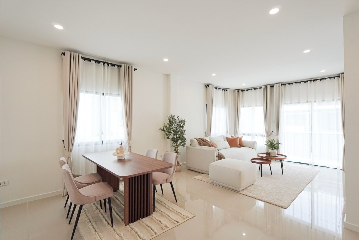 Bright open-plan living and dining room with white walls, beige curtains, a wooden dining table, pink upholstered chairs, a cream sofa, and potted plants.