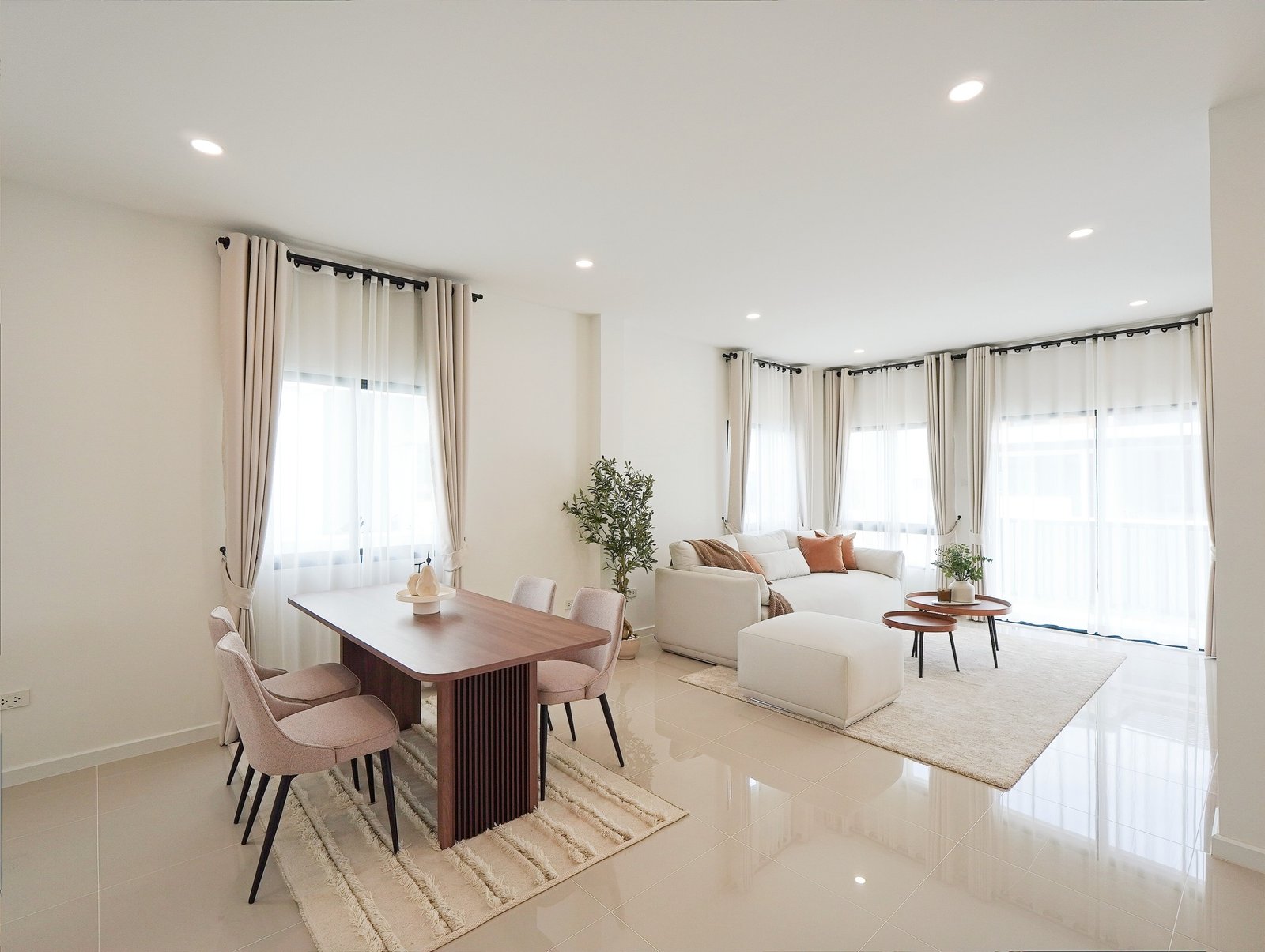 Tropical_Village_3_Pattaya_Dlh453_5 Bright open-plan living and dining room with white walls, beige curtains, a wooden dining table, pink upholstered chairs, a cream sofa, and potted plants.