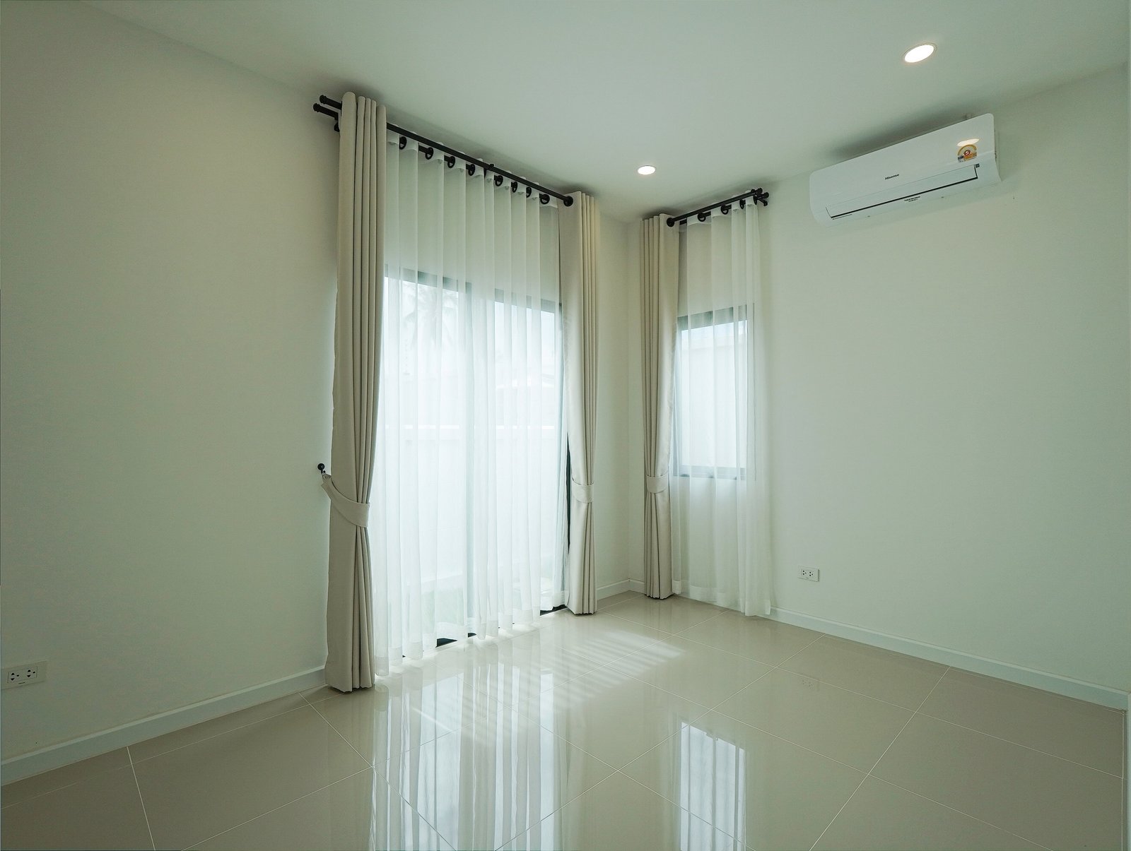 Tropical_Village_3_Pattaya_Dlh453_7 Bright empty room with large windows and sheer white curtains, reflecting light on a glossy tiled floor; wall-mounted air conditioner on the right.