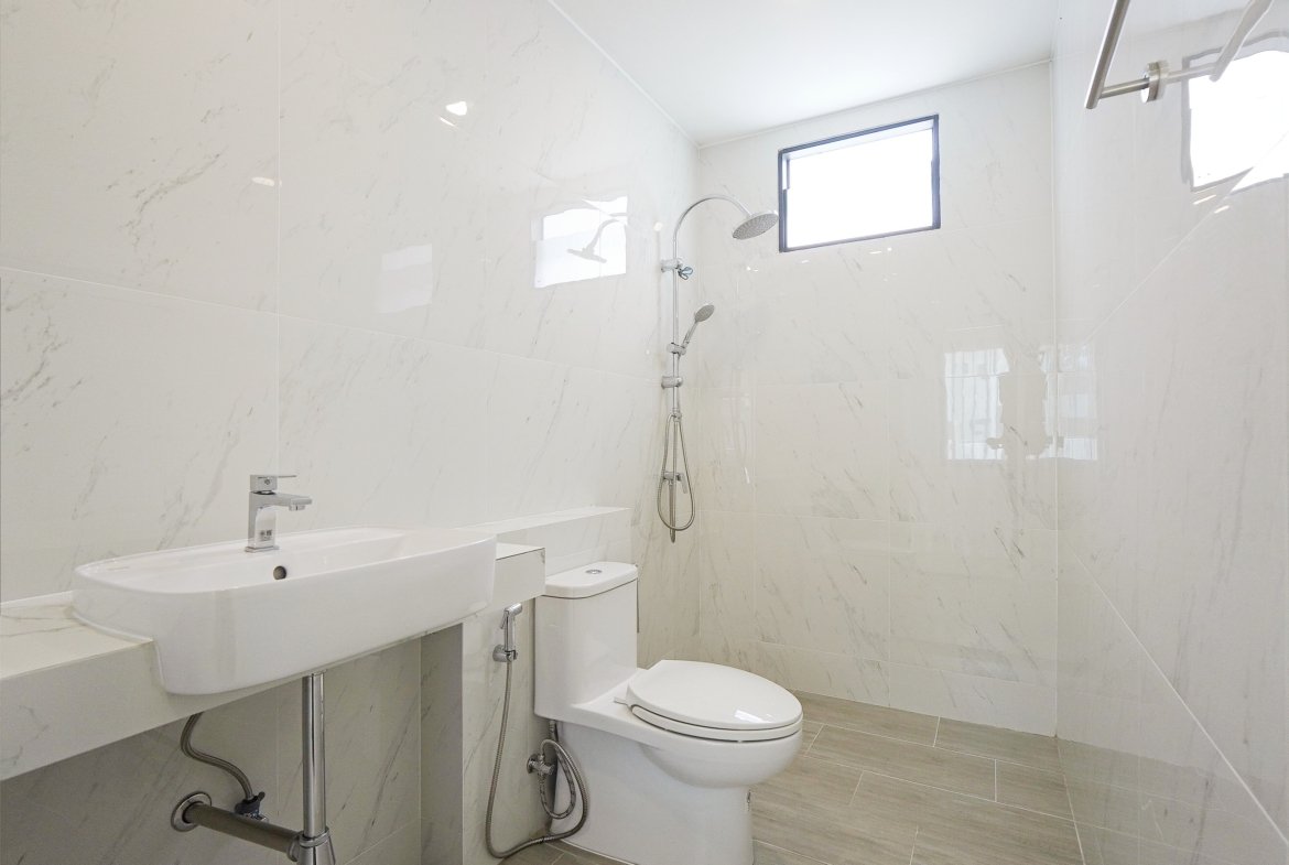 White marble-tiled bathroom with a wall-mounted sink, toilet, and a modern shower area featuring a rainfall head and handheld sprayer, window on the far wall.